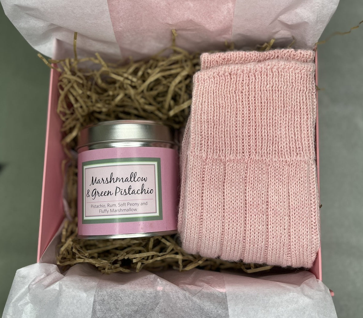 A gift box containing a pink candle and a pale pink Alpaca Bed sock, arranged in shredded paper filler inside a pink box with tissue paper.