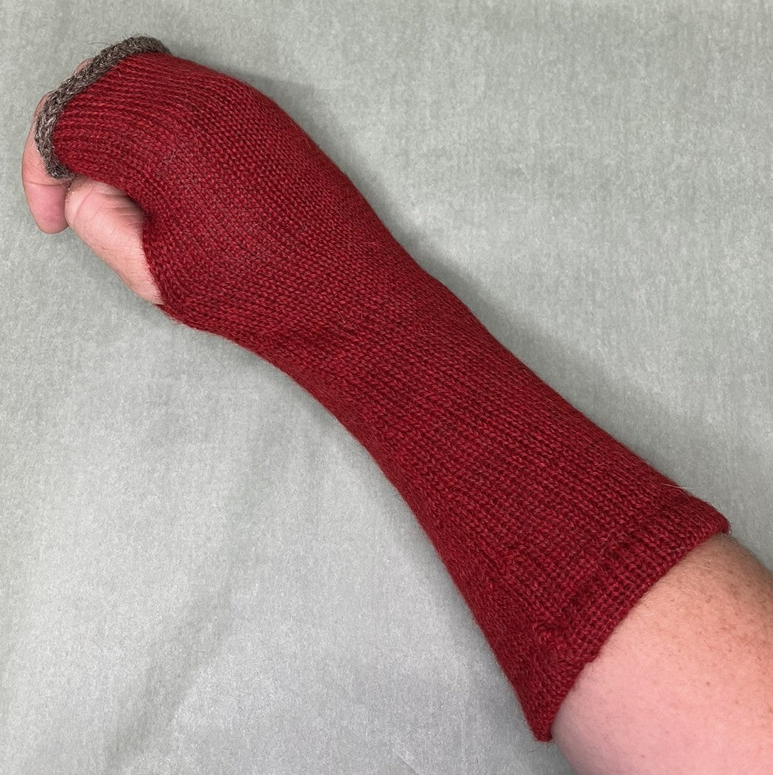 A hand wearing a knitted fingerless glove or wrist protector in deep red, with a textured gray cuff at the top, shown against a light gray fabric background.