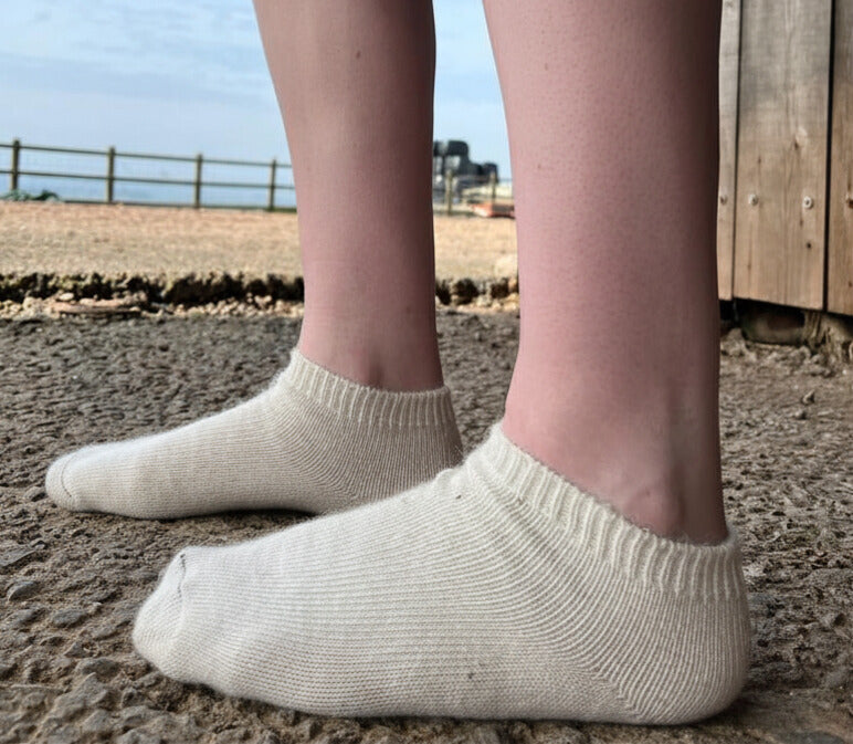 cream alpaca trainer socks worn outdoors on a gravel surface with a wooden structure and blue sky in the background.