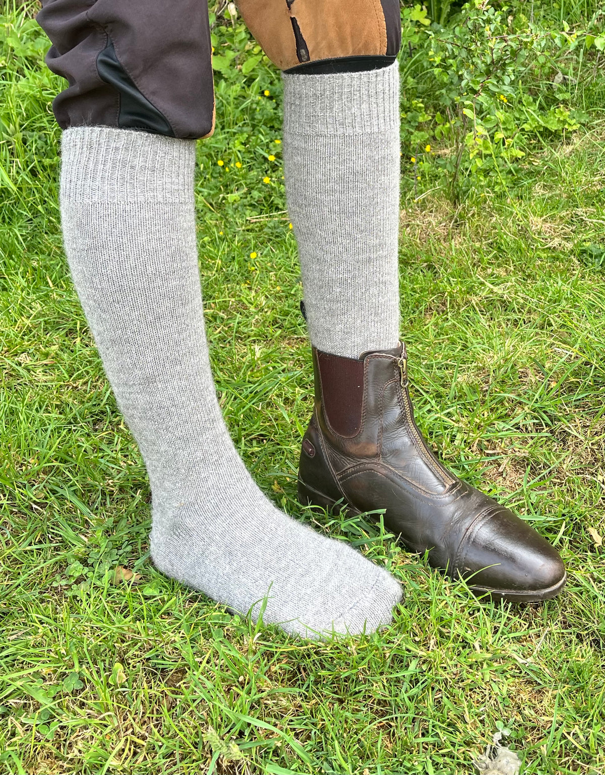Person wearing grey long activity alpaca socks and brown boots on grass