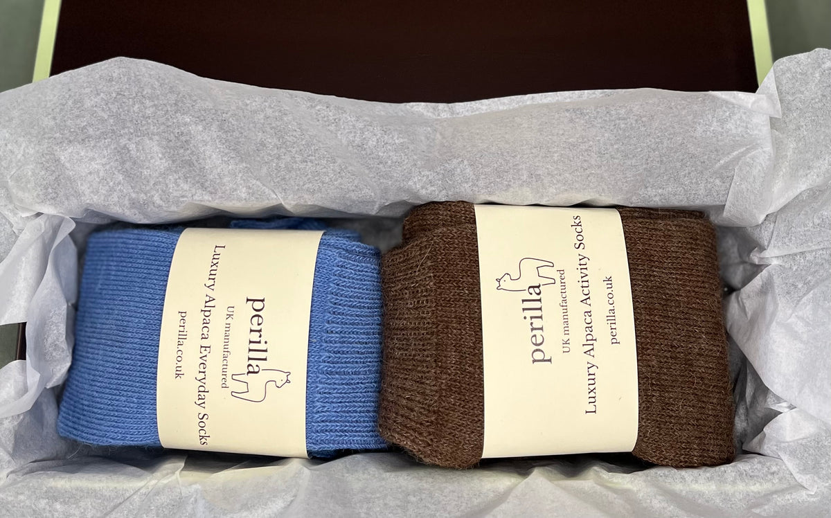 Two pairs of Perilla branded luxury alpaca activity socks, one mid blue and one brown, wrapped in tissue paper inside a gift box.