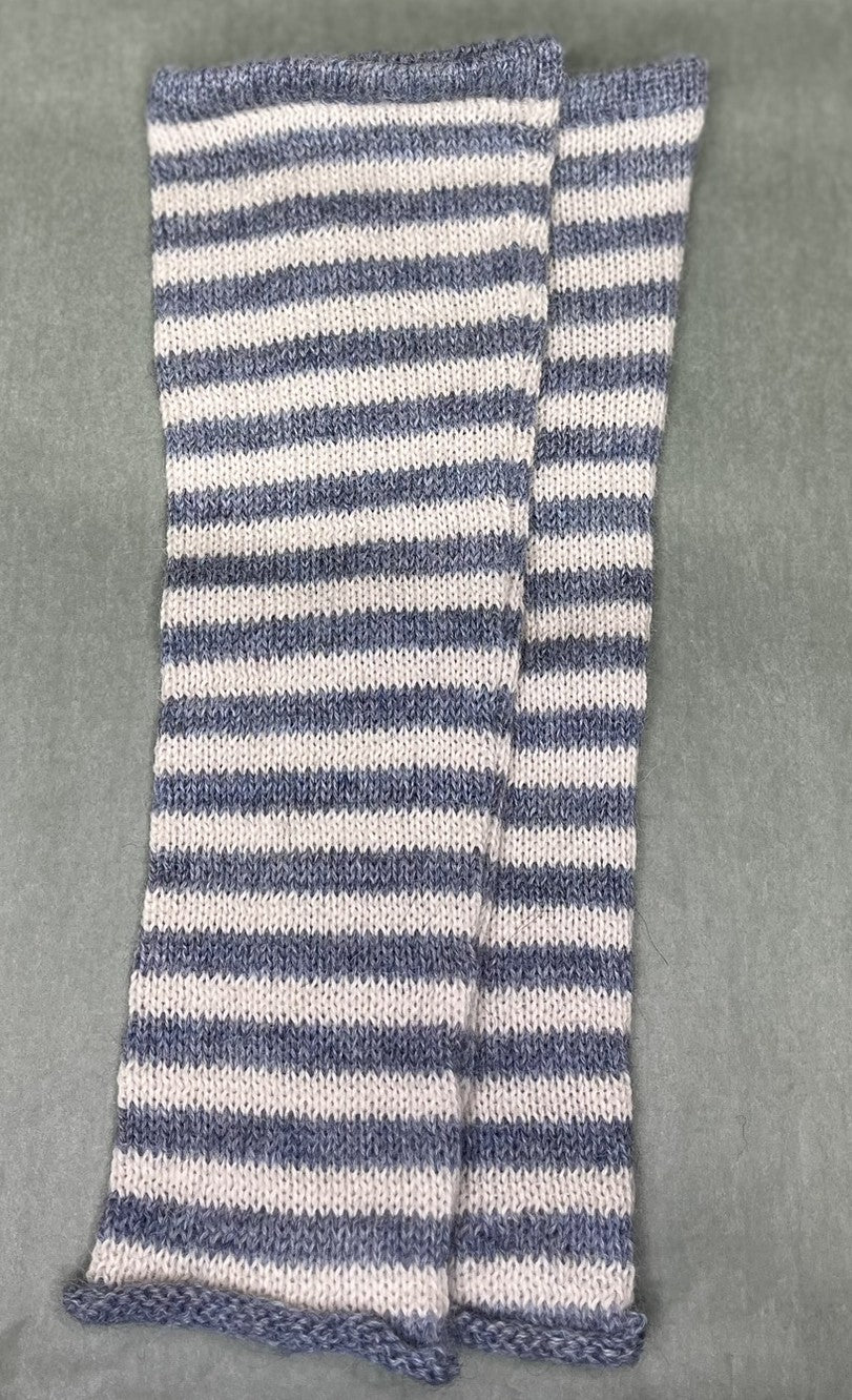 A pair of hand-knit arm warmers featuring horizontal stripes of dusty blue and off-white, laid flat on a textured gray background.