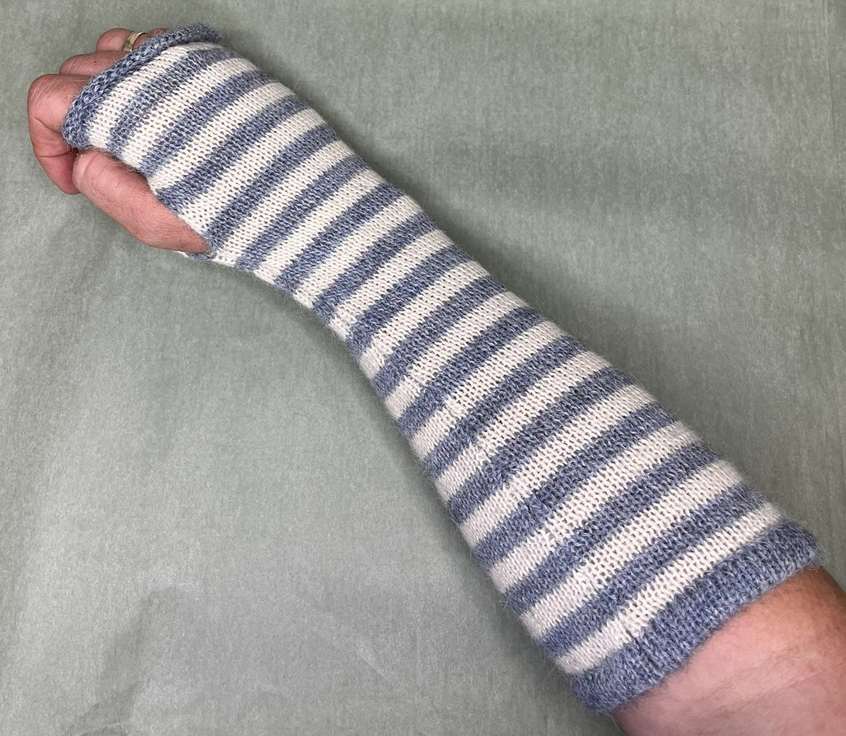 A close-up of a person's forearm and hand wrapped in a knitted arm sleeve with horizontal blue-gray and off-white stripes, set against a textured light gray background.