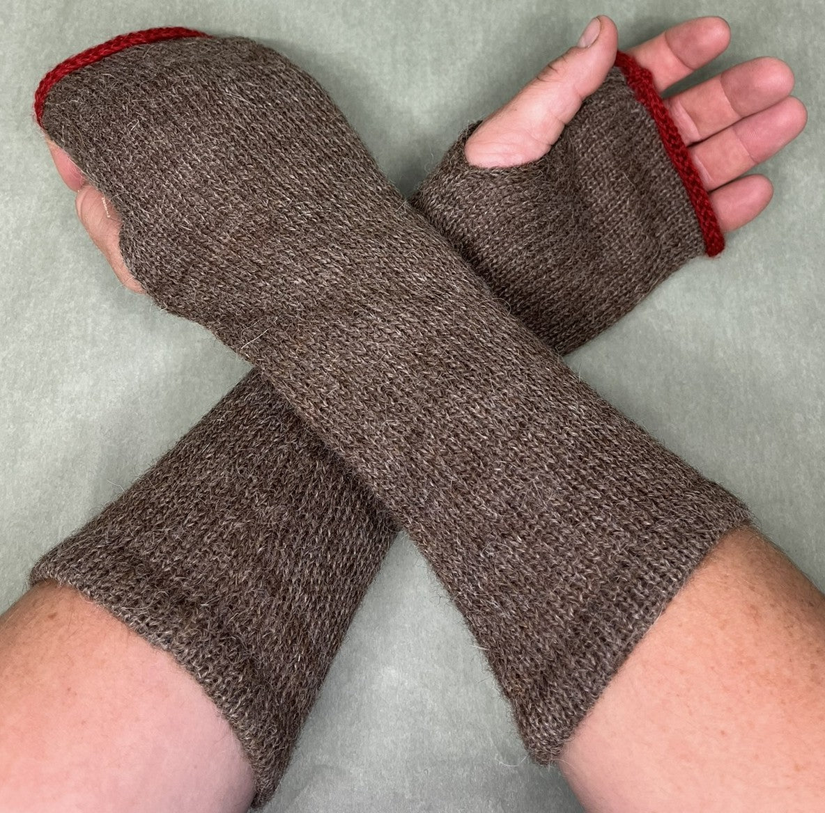 A pair of brown knit fingerless wrist warmers with red trim, shown worn on hands against a light gray background.