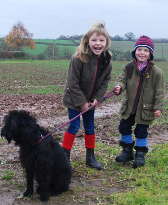 Two children standing outdoors on a muddy field with a black dog, wearing heavy jackets and boots. One child wears a brown jacket with cherry red alpaca socks, and the other wears an olive green jacket with a red and blue striped beanie and mid blue alpaca socks