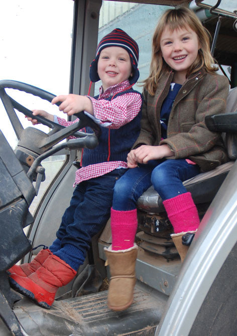 Two children sitting on a vehicle, wearing thick alpaca walking socks in pink and navy colours, with hand-linked toes.