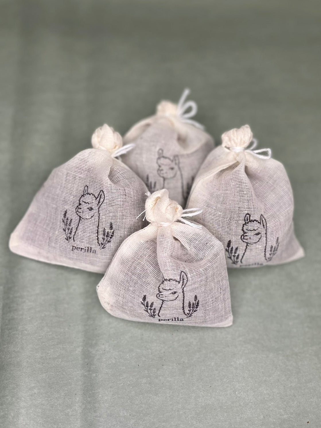 Four small drawstring pouches made of light gray linen fabric, each printed with a black alpaca and perilla 