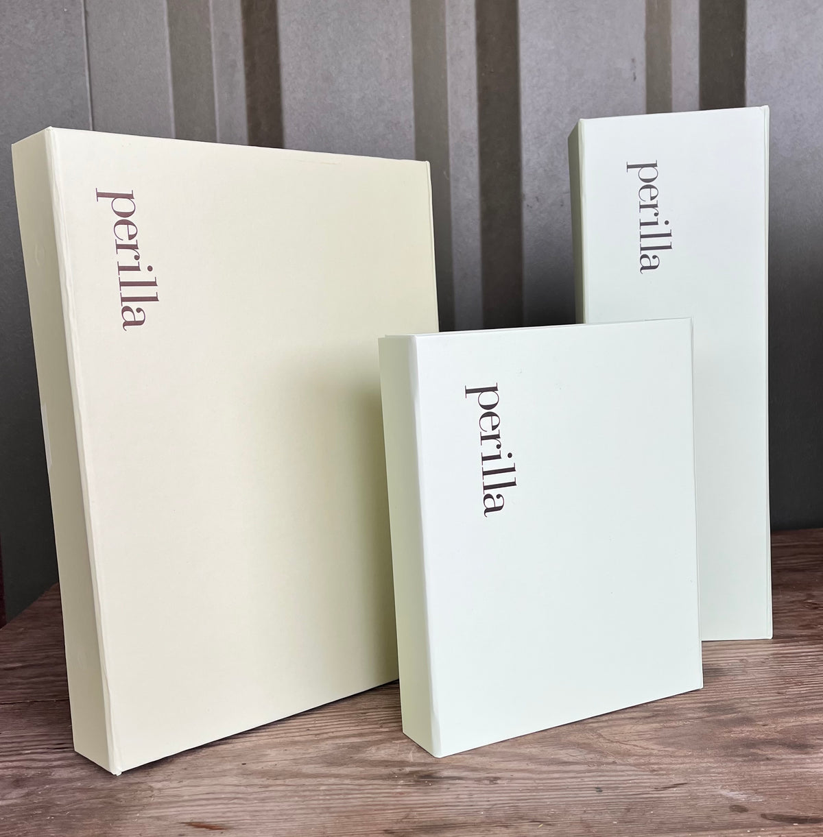 Three Perilla brand product boxes in pale cream and mint green tones arranged on a wooden surface against a gray paneled background.