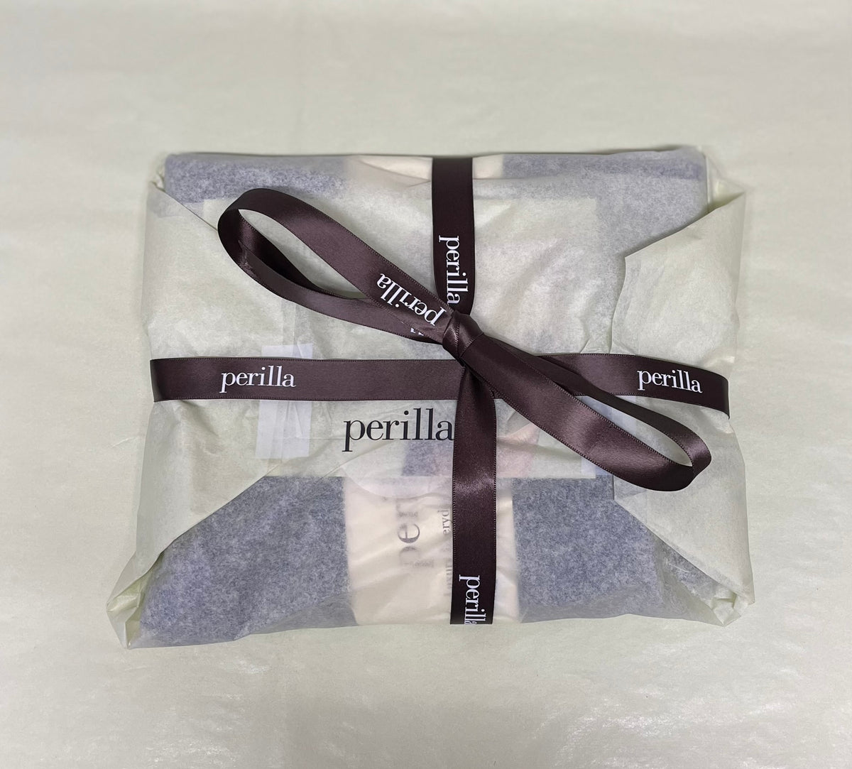 A pair of Perilla alpaca socks wrapped in translucent tissue paper and tied with a brown satin ribbon printed with the Perilla brand name, shown on a light neutral background.