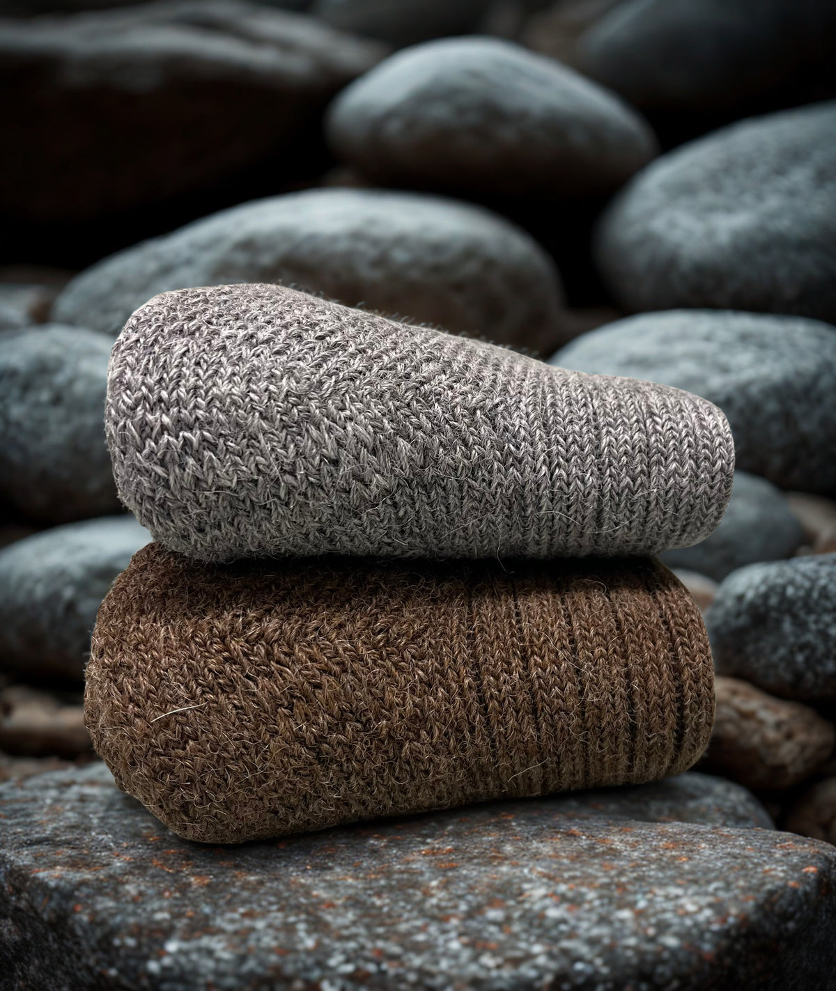 Two folded non-elasticated activity alpaca socks, one grey and one brown, stacked on a stone surface with pebbles in the background.