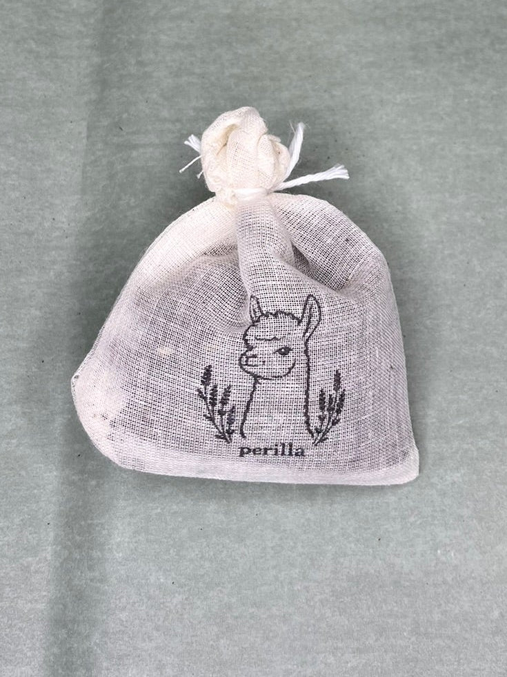 A small lavender linen drawstring pouch with a black alpaca illustration and the word 'perilla' printed on the front, resting on a textured sage green background.