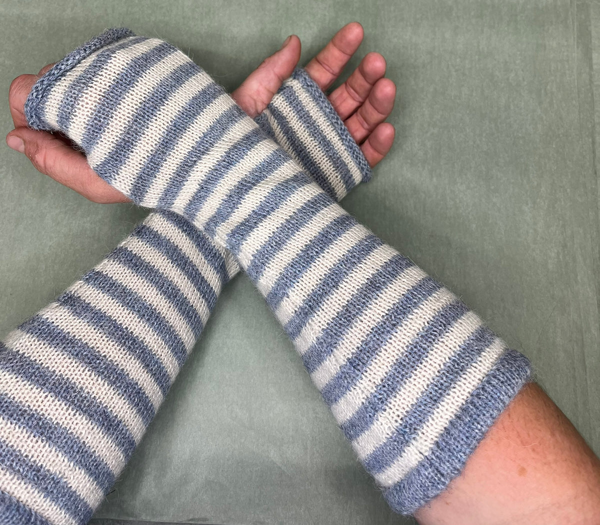 A pair of hand-knit striped arm warmers worn on both forearms, featuring alternating stripes of dusty blue and cream. The hands and forearms are displayed against a solid sage green background.