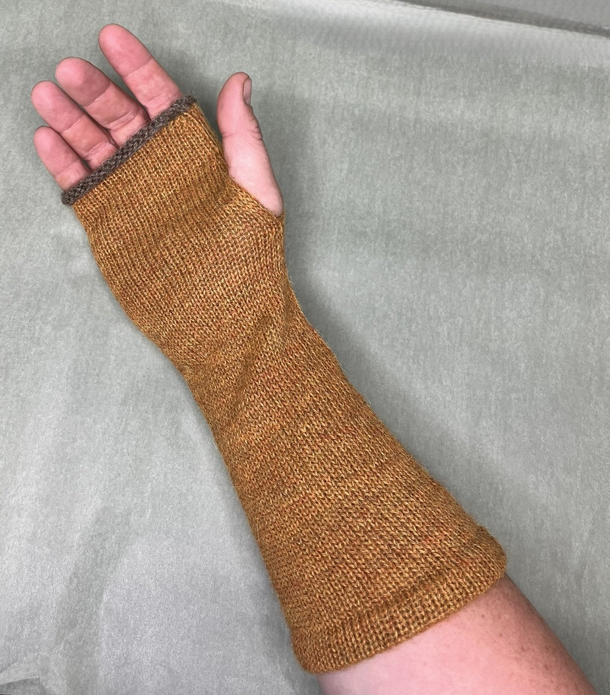 A close-up of a hand wearing a knitted fingerless wrist and forearm sleeve in a warm mustard-brown color, set against a neutral gray background.